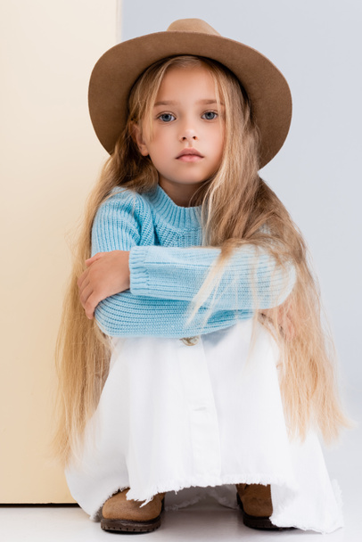 fashionable blonde girl in brown hat and boots, white skirt and blue sweater sitting near beige wall - Photo, Image