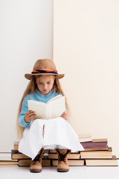 chica rubia de moda en sombrero marrón y botas, falda blanca y suéter azul sentado en libros antiguos y leyendo cerca de la pared beige - Foto, imagen