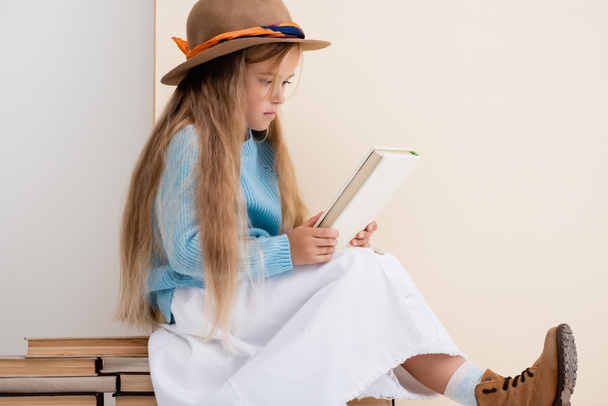 модная блондинка в коричневой шляпе и сапогах, белая юбка и голубой свитер сидят на старинных книгах и читают возле бежевой стены - Фото, изображение
