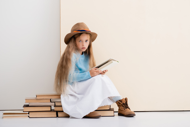 modna blondynka dziewczyna w brązowym kapeluszu i buty, biała spódnica i niebieski sweter siedzi na książkach vintage i czytania w pobliżu beżowej ściany - Zdjęcie, obraz