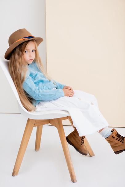 side view of fashionable blonde girl in brown hat and boots, white skirt and blue sweater sitting on chair near beige wall - 写真・画像
