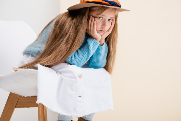 triste chica rubia de moda en sombrero marrón, falda blanca y suéter azul sentado en la silla cerca de la pared beige - Foto, imagen