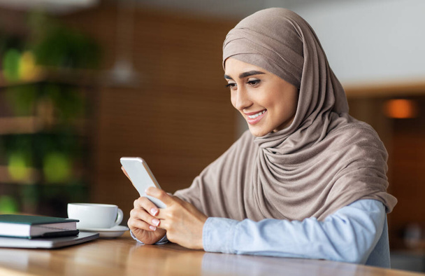 Усміхнена дівчина в хіджабі використовує смартфон під час відпочинку в кафе
 - Фото, зображення
