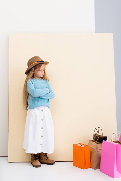 triest modieus blond meisje in bruine hoed en laarzen, witte rok en blauwe trui in de buurt van kleurrijke winkeltassen en beige muur - Foto, afbeelding