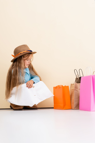 sad fashionable blonde girl in brown hat and boots, white skirt and blue sweater sitting near colorful shopping bags and beige wall - Zdjęcie, obraz