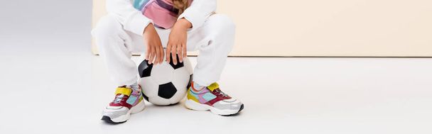 vista ritagliata di ragazza in abbigliamento sportivo in posa con pallone da calcio su sfondo beige e bianco, colpo panoramico - Foto, immagini
