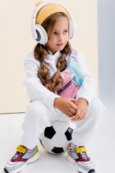 blondýny dívka ve sportovním oblečení a sluchátka pózování s fotbalovým míčem na béžové a bílé pozadí - Fotografie, Obrázek