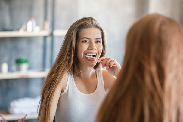 Oral hygiene. Attractive woman cleaning her teeth near looking glass at bathroom - Фото, изображение