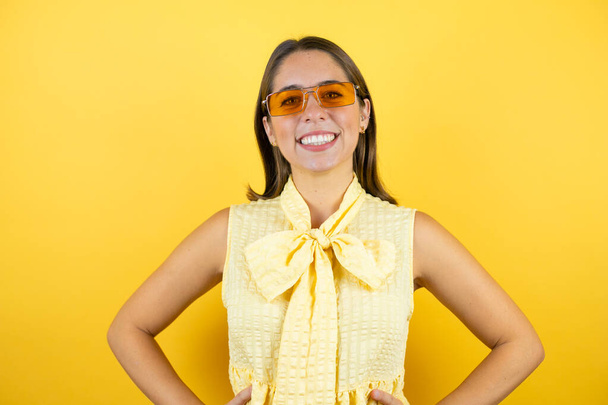 Young beautiful woman wearing sunglasses over isolated yellow background smiling with her arms in her waist - Фото, зображення