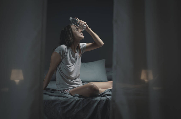 Young woman suffering from insomnia and heat, she is sitting on the bed and holding a water bottle - Φωτογραφία, εικόνα