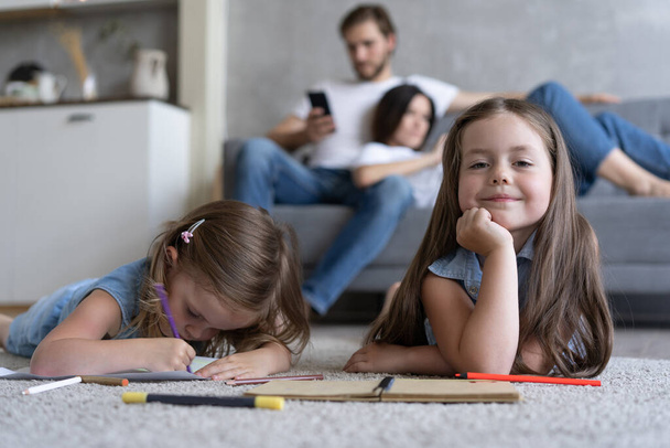 Dzieci siostry bawią się rysunkiem na podłodze podczas gdy młodzi rodzice relaksują się w domu na sofie, małe dziewczynki bawią się - Zdjęcie, obraz