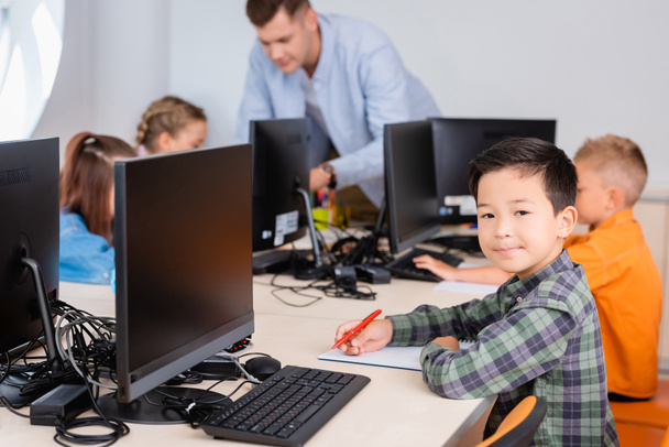 Селективный фокус азиатского школьника, смотрящего в камеру во время записи на ноутбуке рядом с компьютерами в начальной школе  - Фото, изображение