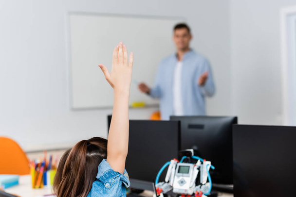 Selective focus of schoolgirl with raised hand near robot and computers in stem school  - Photo, Image