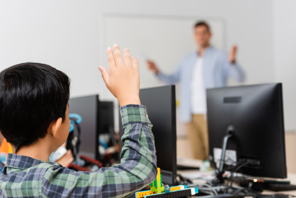 Selective focus of schoolboy with raised hand sitting near robot and computers during lesson in stem school - Фото, изображение