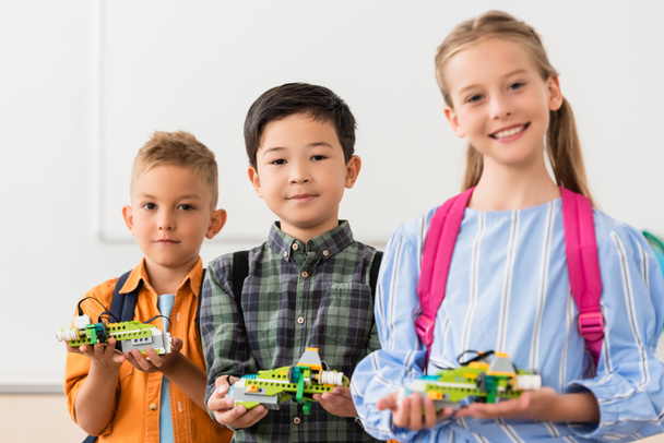 Selective focus of multiethnic pupils holding robots and looking at camera in stem school  - Foto, Bild
