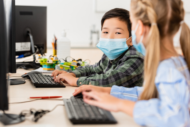 Selective focus of asian schoolboy in medical mask using computer near friend in classroom  - Fotó, kép
