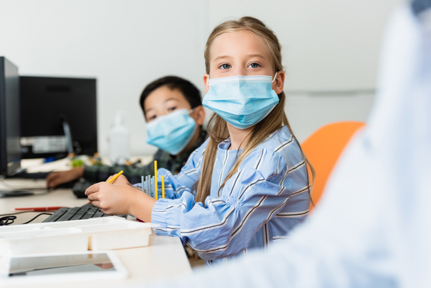 Selective focus of multiethnic pupils in medical masks sitting near computers in stem school  - Photo, Image