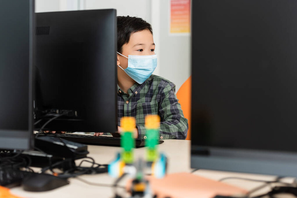 Selective focus of asian schoolboy in medical mask sitting near computers in stem school  - Foto, imagen