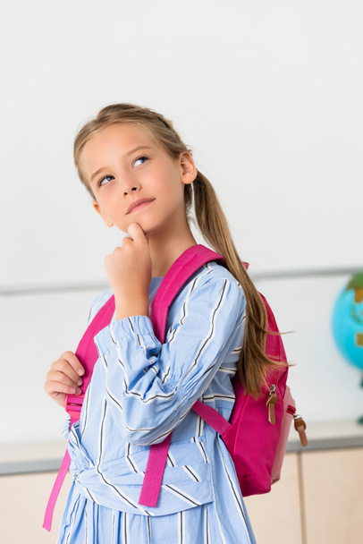 Dreamy schoolgirl with backpack looking away in classroom  - Foto, afbeelding
