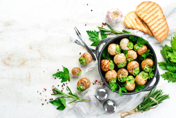 Baked snails with garlic butter and fresh herbs on a black plate on a white wooden background. Top view. Free space for your text. - Foto, immagini