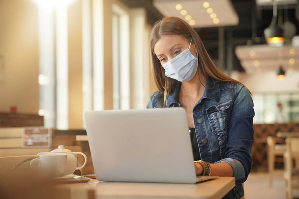 Young woman sitting at coffee shop and working on laptop with face mask - Fotó, kép