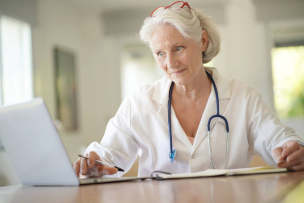 Portrait of smiling senior woman doctor with white hair, working on laptop - Фото, изображение