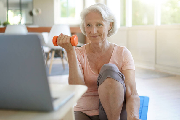 Senior woman doing fitness exercises at home through virtual class - Zdjęcie, obraz