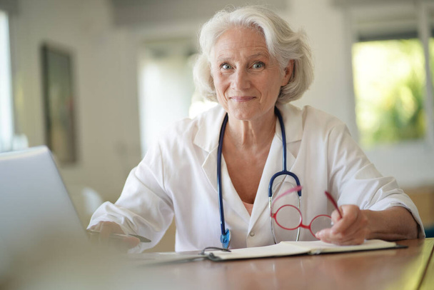 Portrait of smiling senior woman doctor with white hair, working on laptop - Photo, image