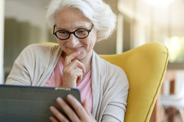 Senior woman relaxing in armchair and using digital tablet - Foto, Bild