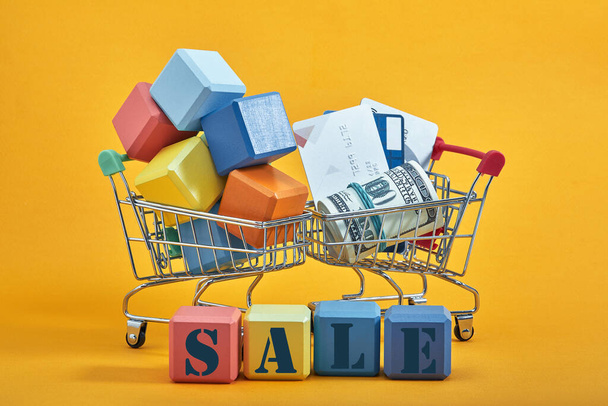 two shopping trolleys with multi-colored cubes, credit cards and dollars on yellow background. Sales word written on cubes. - Fotografie, Obrázek