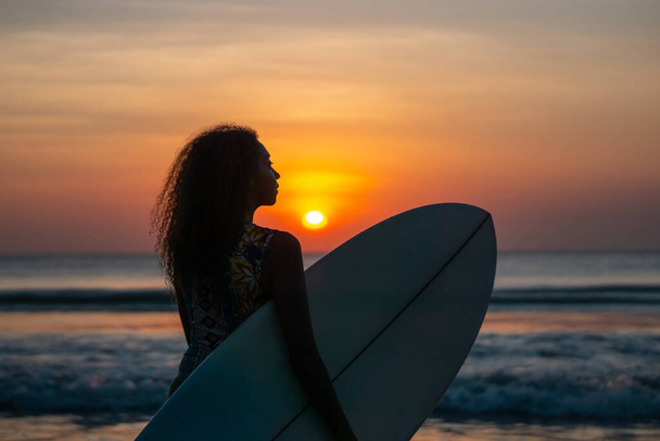 Portrait of woman surfer with beautiful body on the beach with surfboard at colorful sunset in Bali - Foto, Bild