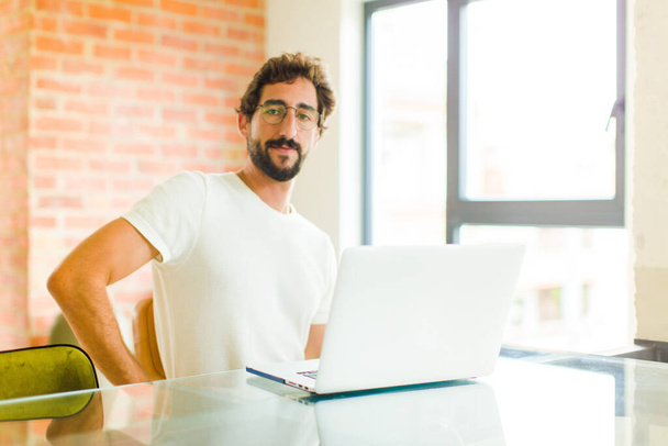 young bearded man with a laptop smiling happily with a hand on hip and confident, positive, proud and friendly attitude - Foto, imagen