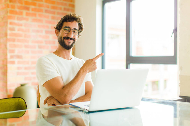 young bearded man with a laptop smiling cheerfully, feeling happy and pointing to the side and upwards, showing object in copy space - Foto, Imagen