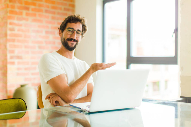 young bearded man with a laptop smiling cheerfully, feeling happy and showing a concept in copy space with palm of hand - Zdjęcie, obraz