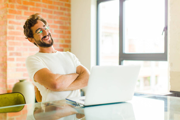 young bearded man with a laptop laughing happily with arms crossed, with a relaxed, positive and satisfied pose - Foto, Bild