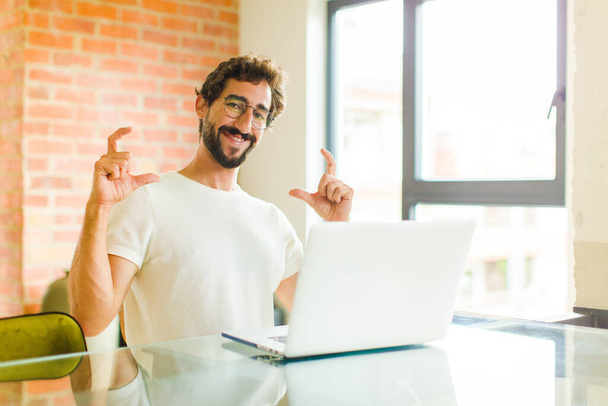 young bearded man with a laptop framing or outlining own smile with both hands, looking positive and happy, wellness concept - Foto, Bild