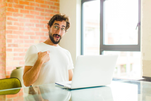young bearded man with a laptop looking happy, proud and surprised, cheerfully pointing to self, feeling confident and lofty - Foto, Imagem
