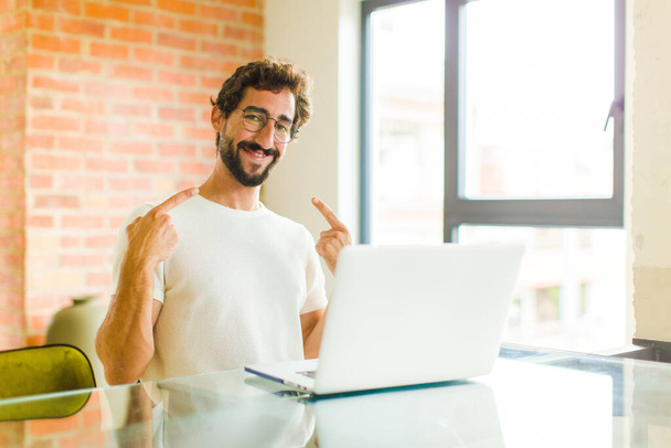 young bearded man with a laptop smiling confidently pointing to own broad smile, positive, relaxed, satisfied attitude - 写真・画像