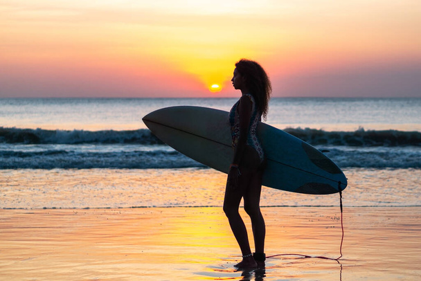 Portrait of woman surfer with beautiful body on the beach with surfboard at colorful sunset in Bali - Фото, изображение