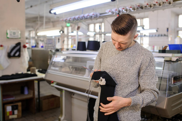 mladý muž analyzující pletený kus látky v blízkosti průmyslových pletených strojů s černou nití v kuželech  - Fotografie, Obrázek