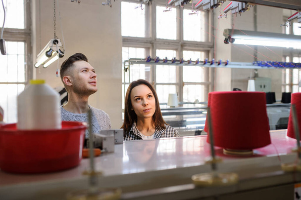zbliżenie zdjęcie młodej kobiety i mężczyzny patrzących i dyskutujących w pobliżu przemysłowej maszyny dziewiarskiej z czerwonymi szyszkami na nim - Zdjęcie, obraz