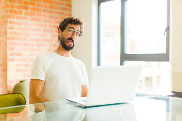 young bearded man with a laptop feeling disgusted and irritated, sticking tongue out, disliking something nasty and yucky - Fotoğraf, Görsel