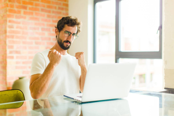 young bearded man with a laptop looking confident, angry, strong and aggressive, with fists ready to fight in boxing position - Foto, imagen