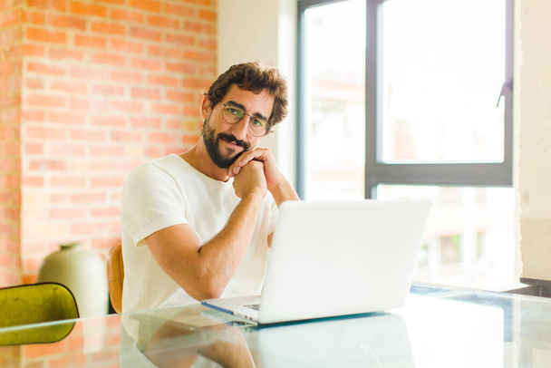 νεαρός γενειοφόρος άνδρας με ένα laptop αίσθημα στην αγάπη και αναζητούν χαριτωμένο, αξιολάτρευτο και ευτυχισμένο, χαμογελώντας ρομαντικά με τα χέρια δίπλα στο πρόσωπο - Φωτογραφία, εικόνα