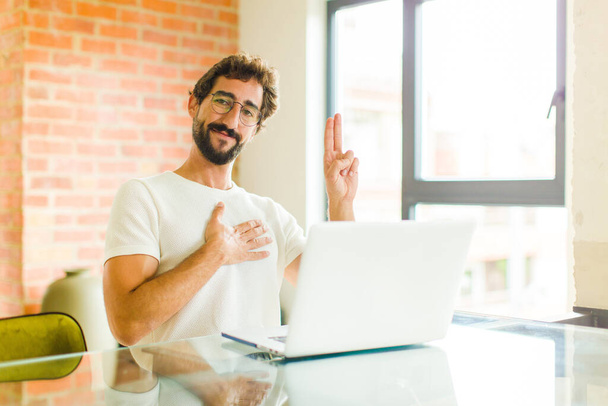 young bearded man with a laptop looking happy, confident and trustworthy, smiling and showing victory sign, with a positive attitude - Foto, afbeelding