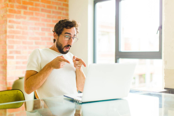 young bearded man with a laptop feeling shocked and surprised, pointing to copy space on the side with amazed, open-mouthed look - 写真・画像