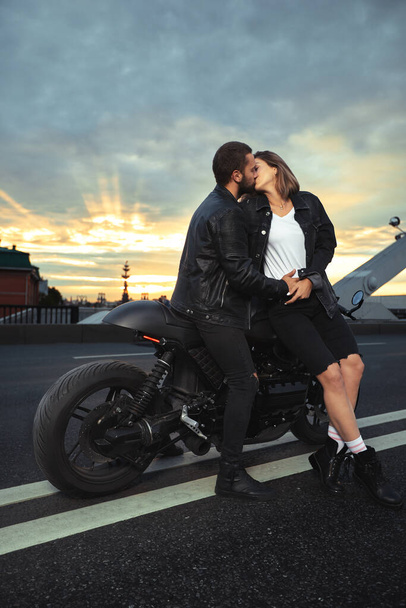 Молодая сексуальная женщина обнимает симпатичного мужчину в стильной черной кожаной куртке, сидит на спортивном мотоцикле на мосту в городе на закате и целуется - Фото, изображение