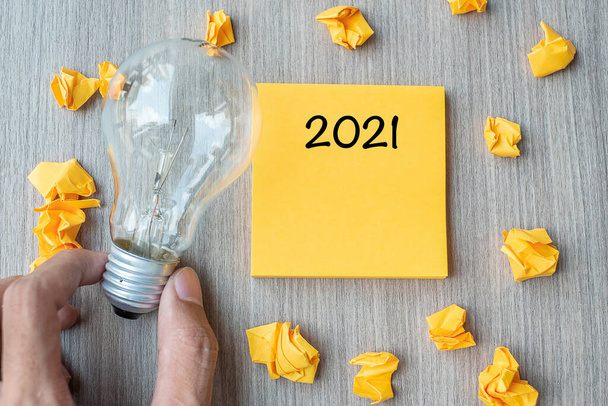 2021 слово на желтой ноте и крошечной бумаге с бизнесменом, держащим лампочку на деревянном фоне стола. Новогодняя идея Креативность, воображение, решимость и целеустремленность - Фото, изображение