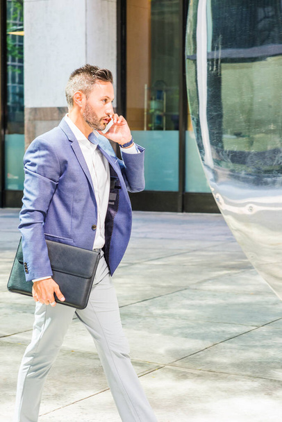 Young European Businessman traveling, working in New York City, with beard, little gray hair, wearing blue jacket, gray pants, holding black leather briefcase, walking on street, talking on cell phone - Foto, Imagen