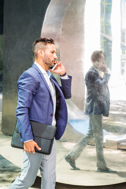 Young European Businessman traveling to work in New York City, with beard, little gray hair, wearing blue jacket, holding black leather briefcase, walking pass by mirror, talking on cell phone - Foto, Imagen